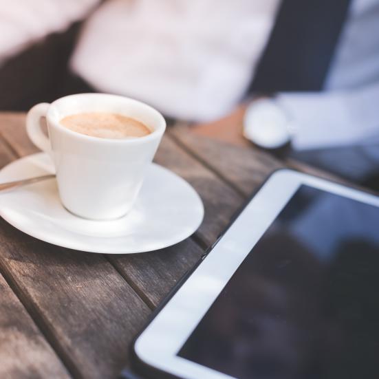 En tablet ligger på et bord og ved siden af står en kop kaffe